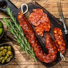 Chorizo co to? Pikantna, wędzona kiełbasa pokrojona w plastry, podana z oliwkami, kaparami i rozmarynem na drewnianej desce.