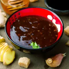 Sos teriyaki przepis: miseczka z ciemnym sosem, obok imbir, czosnek, brązowy cukier i miód.
