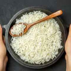 Dwie dłonie trzymają okrągłą patelnię z parującym ryżem basmati. Drewniana łyżka zanurzona w ryżu.