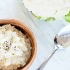 Pyszne puree z kalafiora w drewnianej misce, posypane pieprzem. Obok leży łyżka, a w tle świeży kalafior.