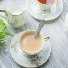 Elegancka filiżanka z herbatą z mlekiem, ozdobiona złotymi wzorami, obok dzbanuszka i czajniczka.