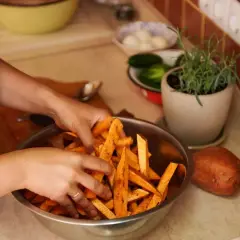 Ręce mieszają pokrojone bataty w misce, przygotowując je do pieczenia. Obok leży cały batat, a w tle zioła i warzywa. Idealne do sosu belgijskiego.