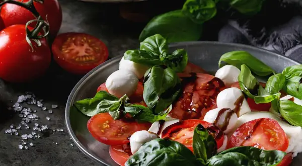 Sałatka caprese: soczyste pomidory, białe kulki mozzarelli i świeża bazylia, polane ciemnym sosem.