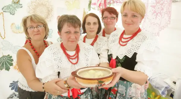 Pięć kobiet w tradycyjnych strojach ludowych prezentuje miskę z zupą. Jedna z nich trzyma ją w dłoniach. W tle widać ozdobne koronki, niczym z kukiełkowego teatru.