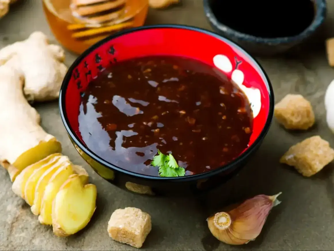 Sos teriyaki przepis: miseczka z ciemnym sosem, obok imbir, czosnek, brązowy cukier i miód.