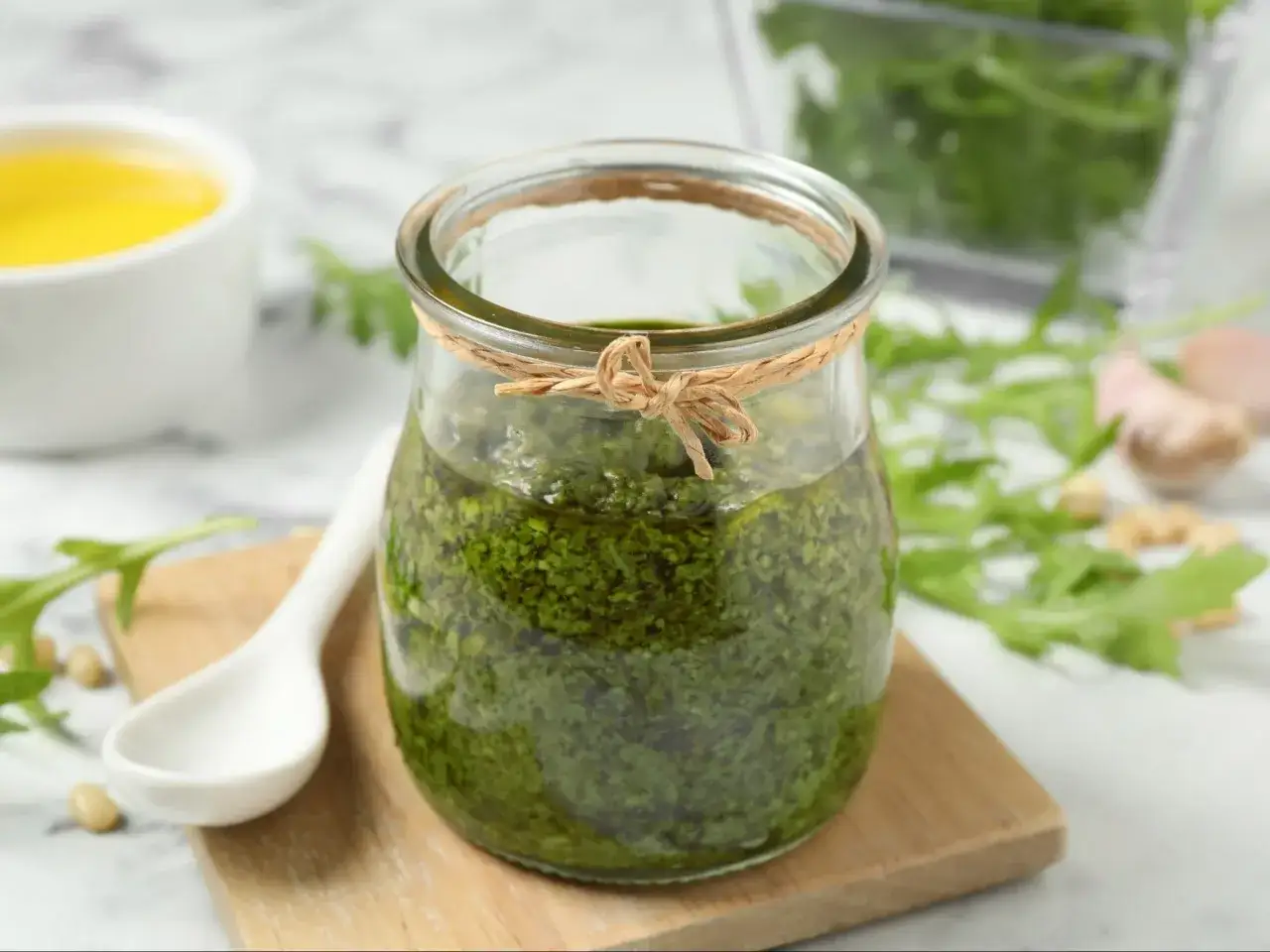Słoik z domowym pesto z rukoli, obok biała łyżeczka, orzeszki piniowe i oliwa.