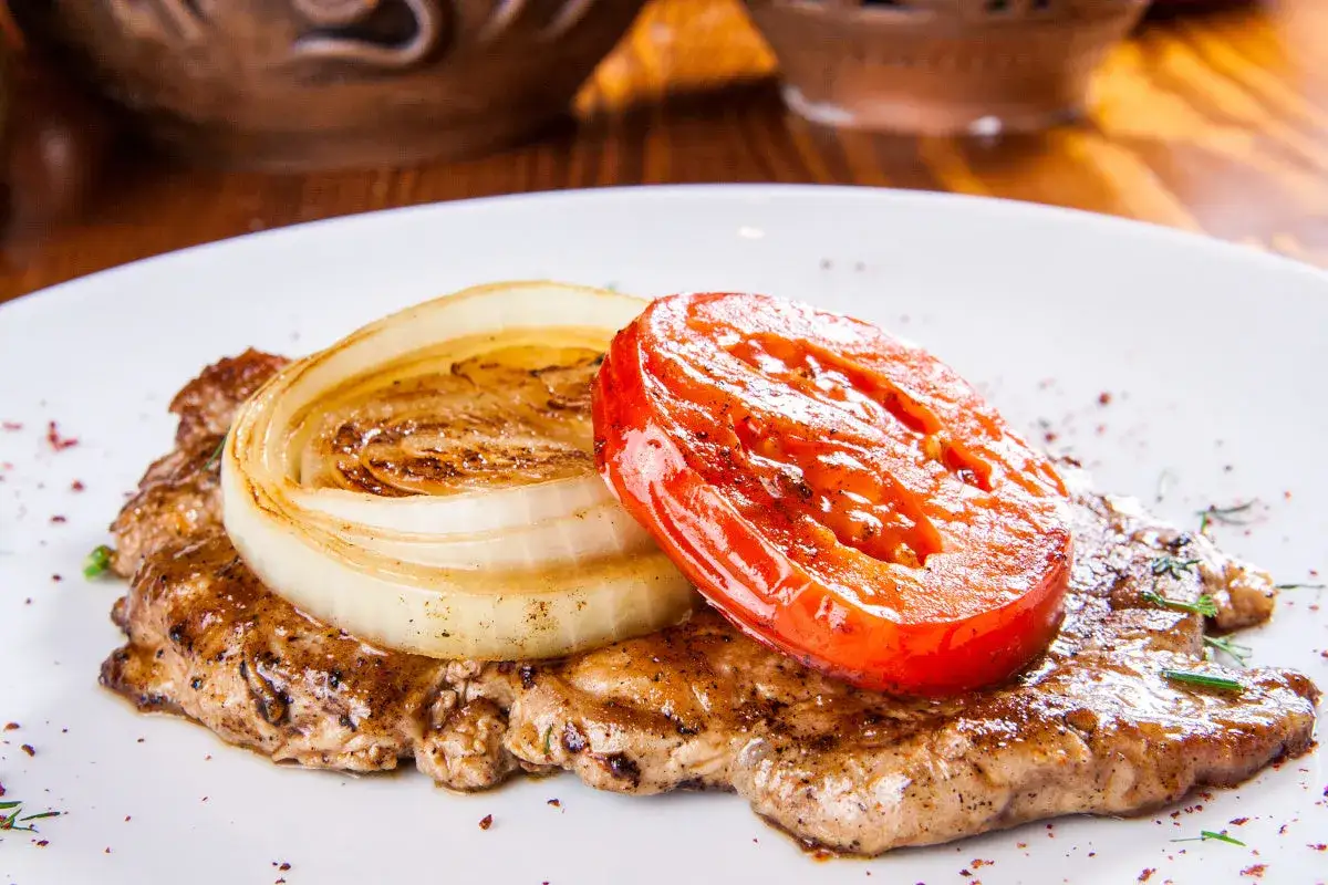 Soczysta karkówka z piekarnika w plastrach, podana z grillowaną cebulą i pomidorem. Pyszne danie na obiad.