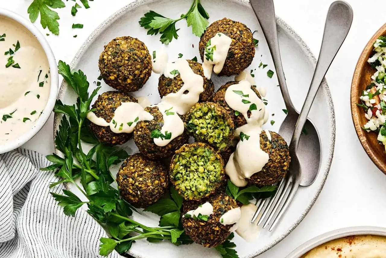 Falafel co to? Aromatyczne kulki z ciecierzycy, polane sosem tahini i posypane natką pietruszki, na talerzu z dodatkami.