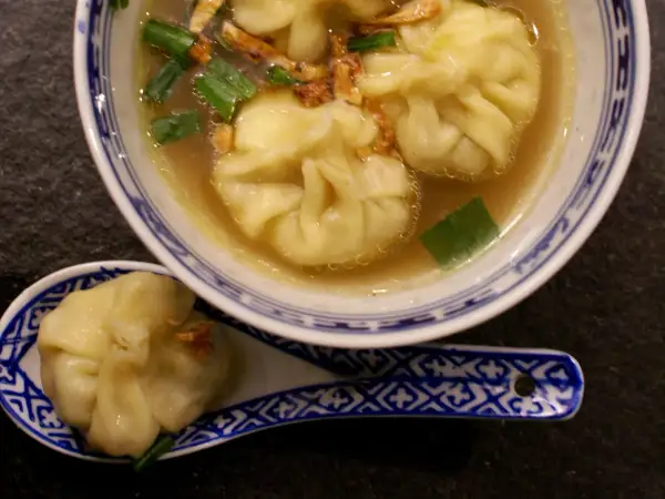 A miseczce z niebiesko-białym wzorem znajduje się aromatyczna zupa wonton z zieloną cebulką i chrupiącymi dodatkami. Na łyżce leży jeden pierożek wonton.