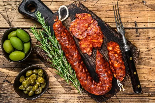 Chorizo co to? Pikantna, wędzona kiełbasa pokrojona w plastry, podana z oliwkami, kaparami i rozmarynem na drewnianej desce.