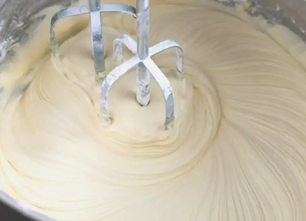 Mieszadła miksera obracają się w gładkim, kremowym **kremie z mascarpone i białej czekolady**, tworząc wiry.