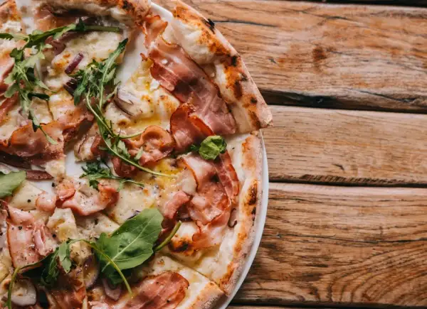 Pyszna pizza carbonara z chrupiącym boczkiem, rukolą i cebulą na drewnianym blacie.