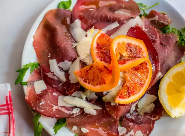 Bresaola co to? Cienkie plastry wędzonej wołowiny, parmezan, rukola i soczyste pomarańcze na talerzu.