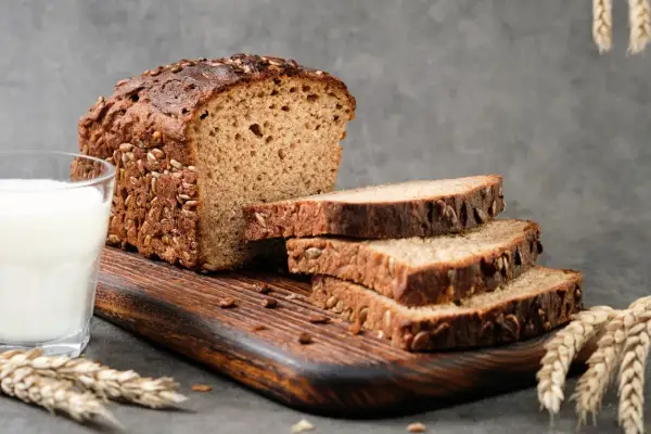 Chleb proteinowy z ziarnami słonecznika i dyni, pokrojony na kromki, obok szklanka mleka i kłosy zboża.