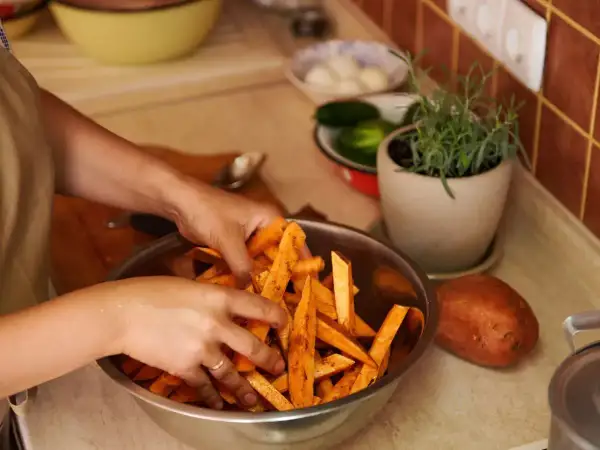 Ręce mieszają pokrojone bataty w misce, przygotowując je do pieczenia. Obok leży cały batat, a w tle zioła i warzywa. Idealne do sosu belgijskiego.