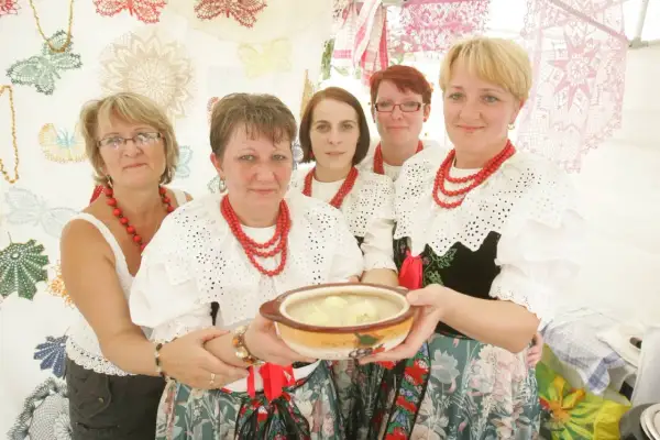 Pięć kobiet w tradycyjnych strojach ludowych prezentuje miskę z zupą. Jedna z nich trzyma ją w dłoniach. W tle widać ozdobne koronki, niczym z kukiełkowego teatru.