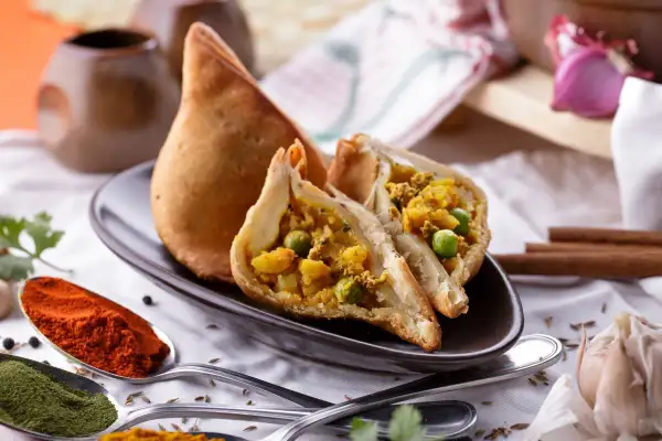 Chrupiący indyjski pierożek samosa, przekrojony na pół, ukazuje bogate nadzienie z ziemniaków i groszku. Obok łyżki z papryką i zielonymi przyprawami.